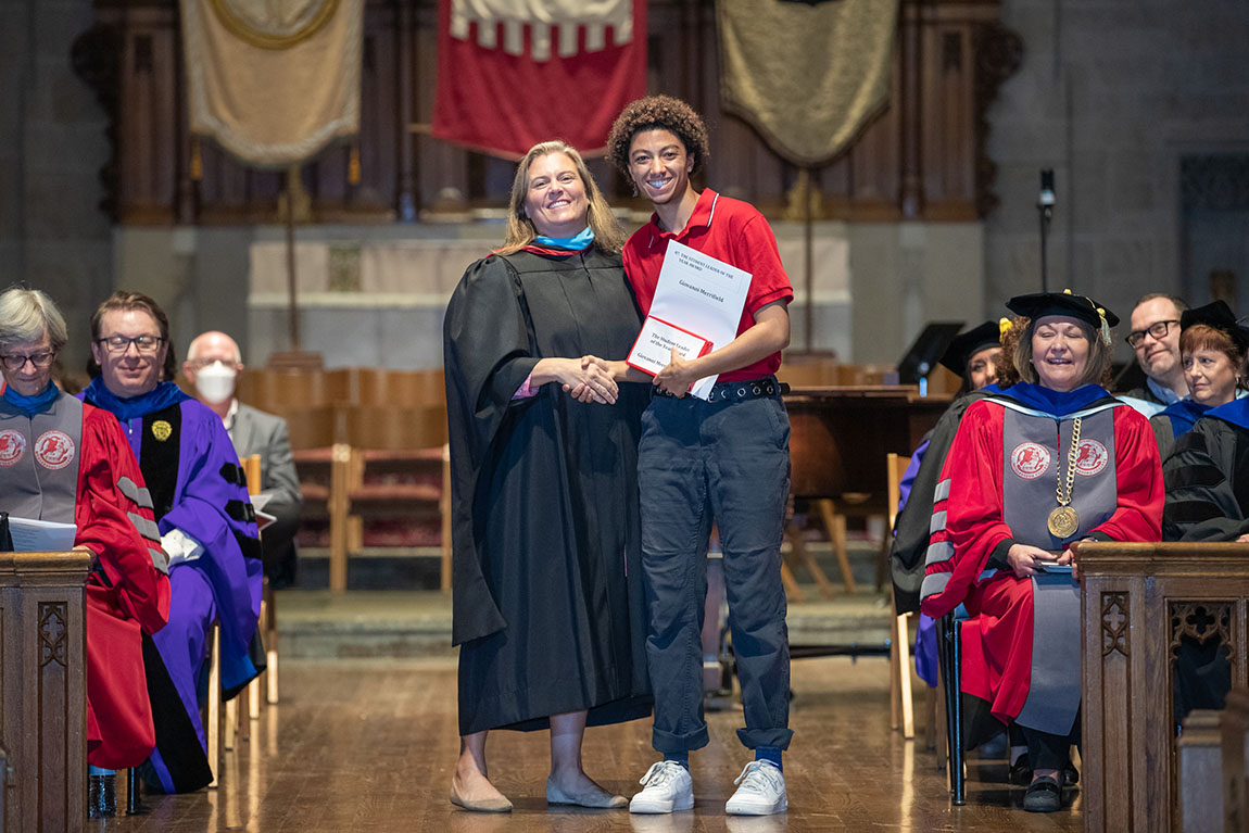 A woman in black academic robes smiles next to a college student in a red shirt and dark pants holding a certificate